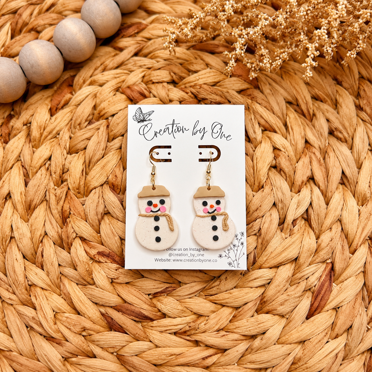 Handmade polymer clay snowman dangle earrings displayed on a branded jewelry card over a woven boho straw background with natural wooden beads and dried flowers. Cute Christmas earrings with gold hooks, rosy cheeks, and a rustic scarf design.