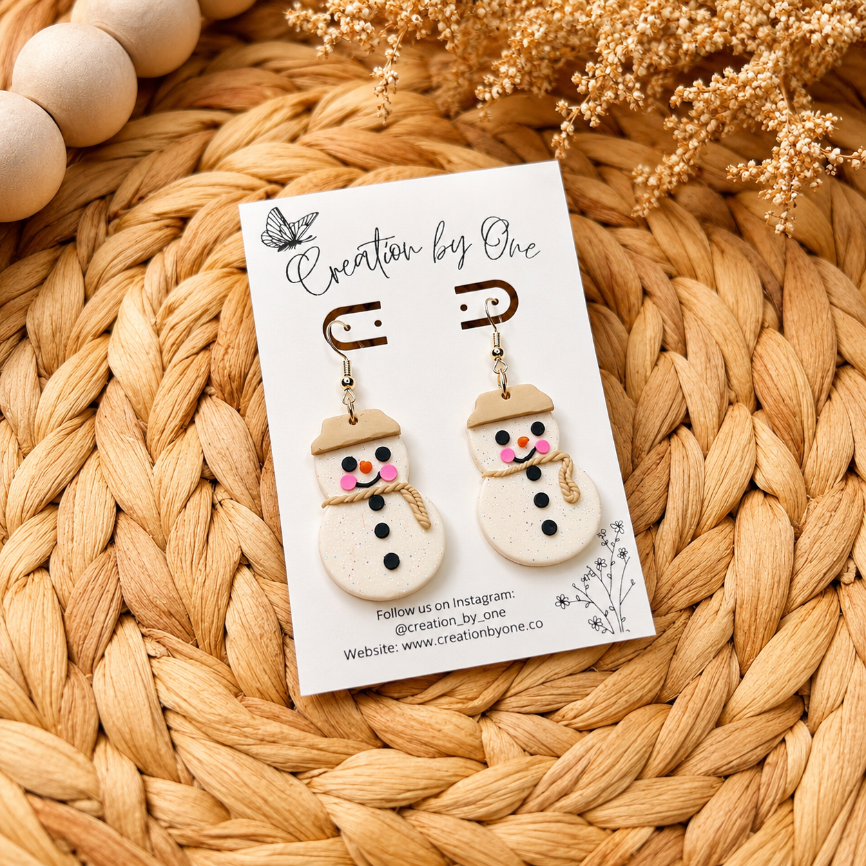 Handmade polymer clay snowman dangle earrings displayed on a branded jewelry card over a woven boho straw background with natural wooden beads and dried flowers. Cute Christmas earrings with gold hooks, rosy cheeks, and a rustic scarf design.