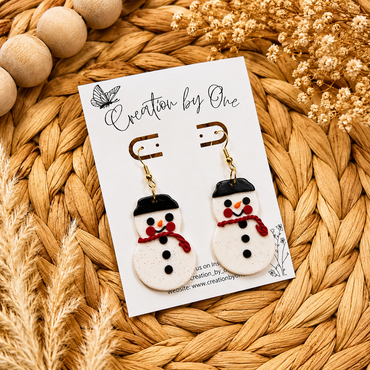 Handmade glitter snowman dangle earrings with red scarves and black hats displayed on a Creation by One jewelry card on a neutral boho woven background with natural dried florals. Festive lightweight polymer clay Christmas earrings perfect for holiday outfits, gifts, and winter accessories.