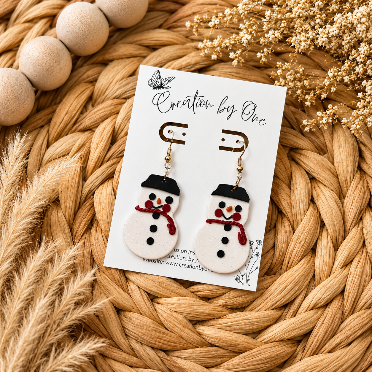 Handmade glitter snowman dangle earrings with red scarves and black hats displayed on a Creation by One jewelry card on a neutral boho woven background with natural dried florals. Festive lightweight polymer clay Christmas earrings perfect for holiday outfits, gifts, and winter accessories.