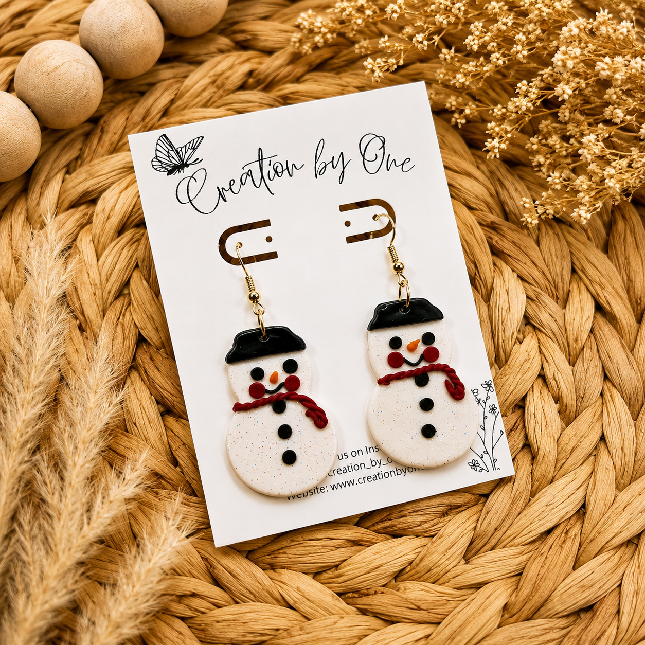 Handmade glitter snowman dangle earrings with red scarves and black hats displayed on a Creation by One jewelry card on a neutral boho woven background with natural dried florals. Festive lightweight polymer clay Christmas earrings perfect for holiday outfits, gifts, and winter accessories.