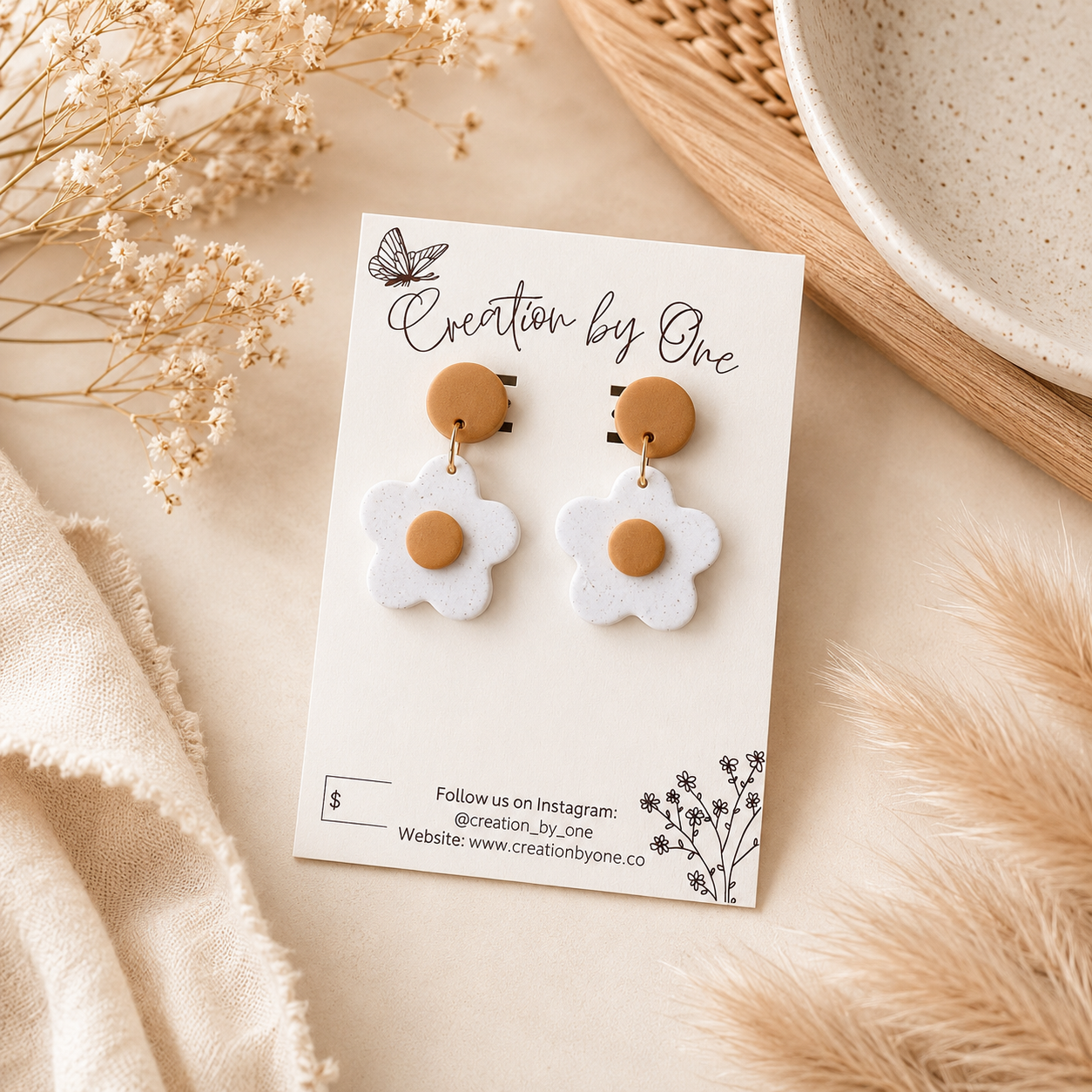 A pair of daisy-shaped drop earrings with white petals and tan centers, displayed on a white card labeled 'Creation by One' in a styled flat-lay with dried flowers and neutral textiles.