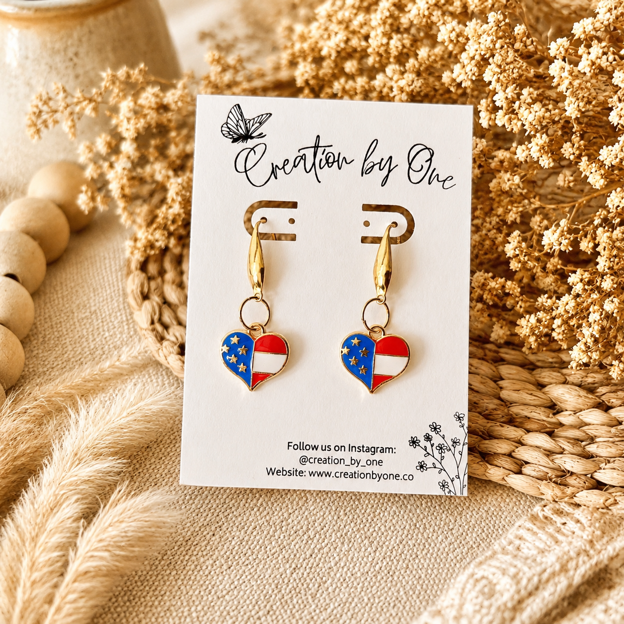Heart-shaped earrings with American flag design on a 'Creation by One' branded card, surrounded by dried flowers and wooden beads.