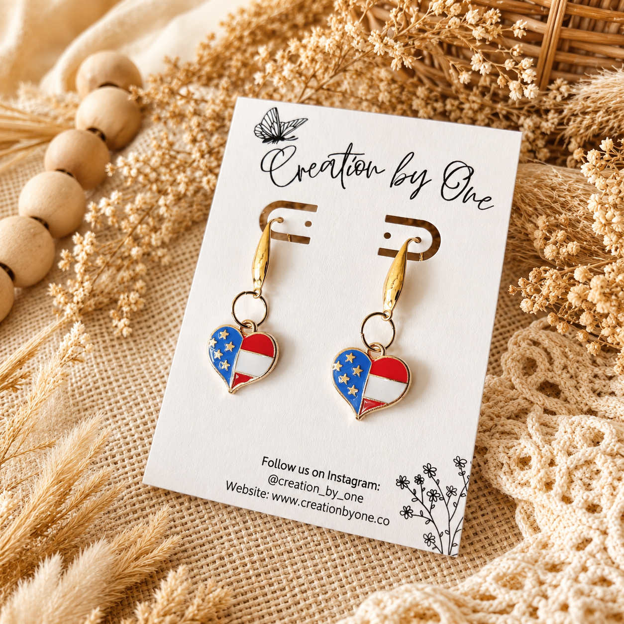 Heart-shaped flag earrings on a 'Creation by One' branded card with dried plants in the background.