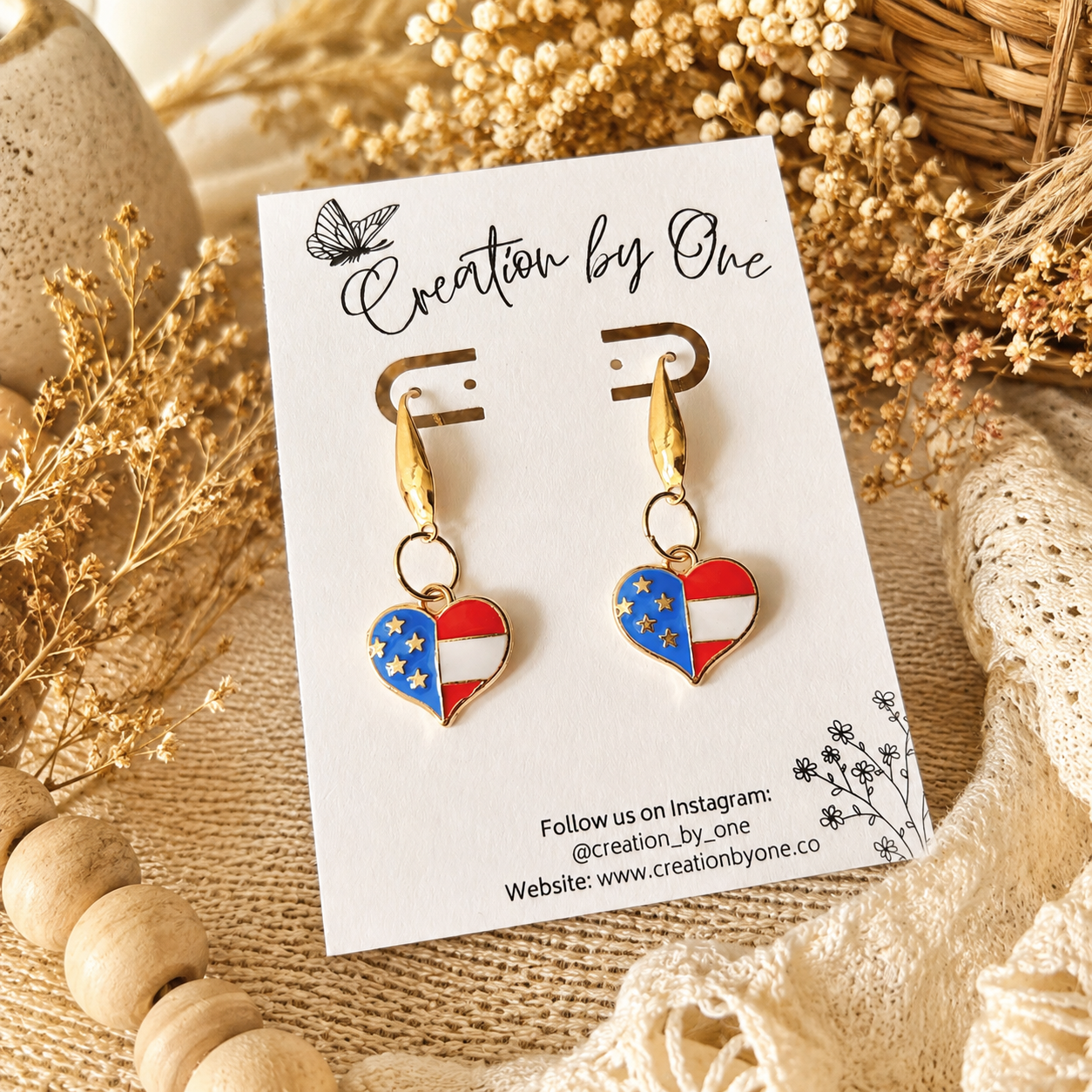 Heart-shaped earrings with American flag design on a 'Creation by One' branded card, surrounded by natural elements.