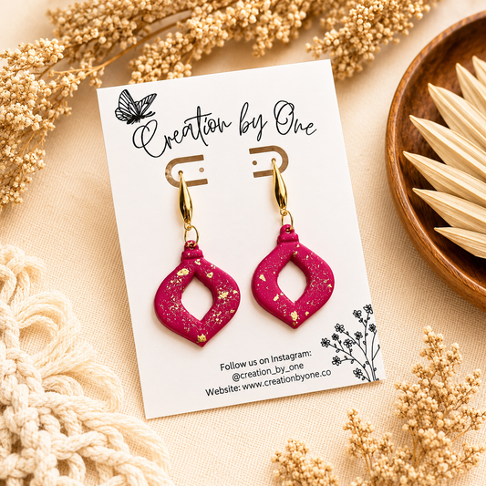 Handmade hot pink polymer clay dangle earrings with gold leaf flakes and gold-tone teardrop studs, displayed on a Creation by One jewelry card against a neutral boho linen background with dried florals.