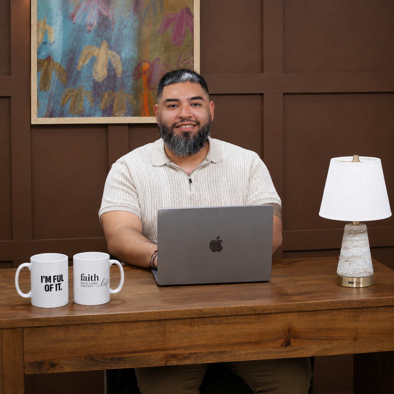 Man sitting at a desk with a laptop, two mugs, and a lamp in a room with a painting on the wall.