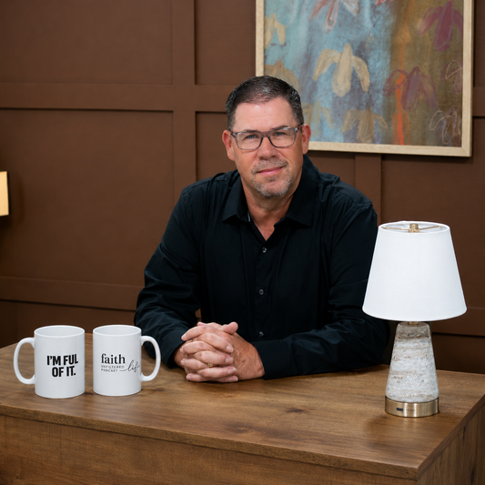 Man sitting at a table with two mugs and a lamp, against a brown wall with abstract art.
