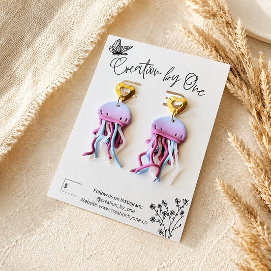 A pair of jellyfish drop earrings displayed on a white branded card, photographed on a beige fabric surface with dried wheat stems around it. The earrings have pastel purple and pink bodies with blue and white trailing strands and gold heart-shaped tops.