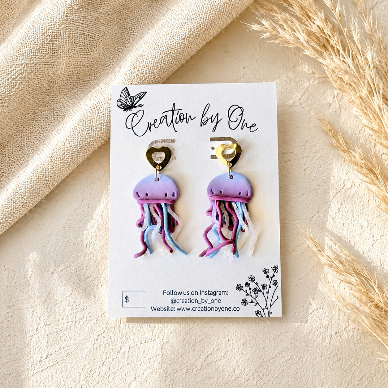 A pair of jellyfish-shaped drop earrings displayed on a white branded card, photographed in a styled flat-lay with beige fabric and dried grass around it. The earrings have pastel purple and pink bodies with blue and pink tentacles and gold heart-shaped hooks.