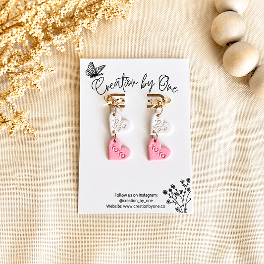Pair of earrings on a card with 'Creation by One' branding, surrounded by decorative elements on a beige surface.