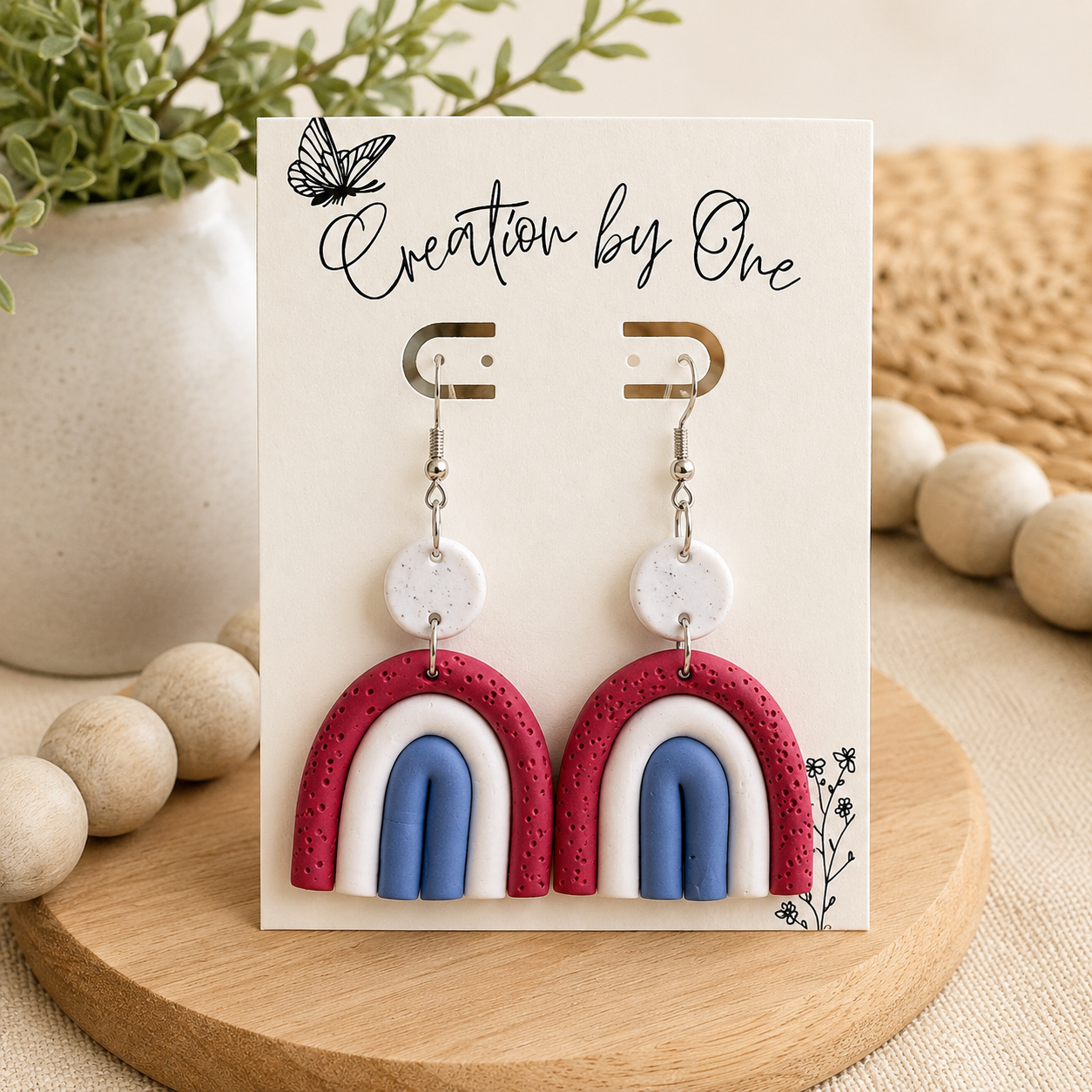 Pair of earrings on a display card with 'Creation by One' branding, set against a natural background.
