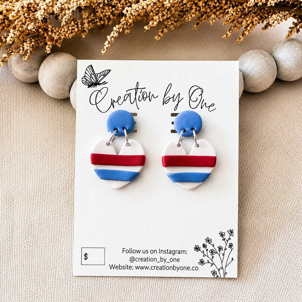 Heart-shaped earrings with blue, red, and white stripes on a 'Creation by One' branded card.