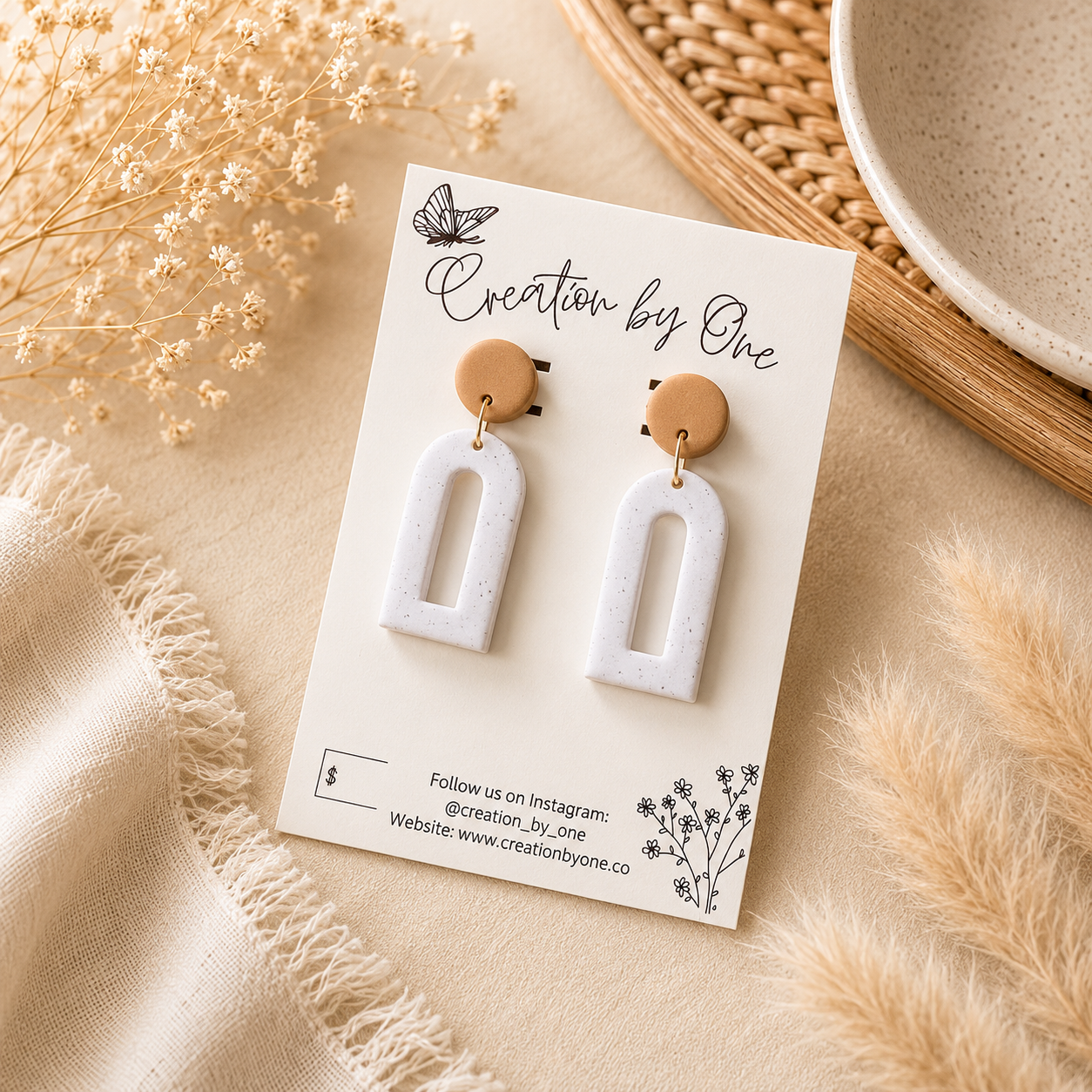 A pair of white arch-shaped drop earrings with round tan wooden studs, displayed on a white card labeled 'Creation by One' in a styled flat-lay setting with beige fabric, dried flowers, and a woven tray.