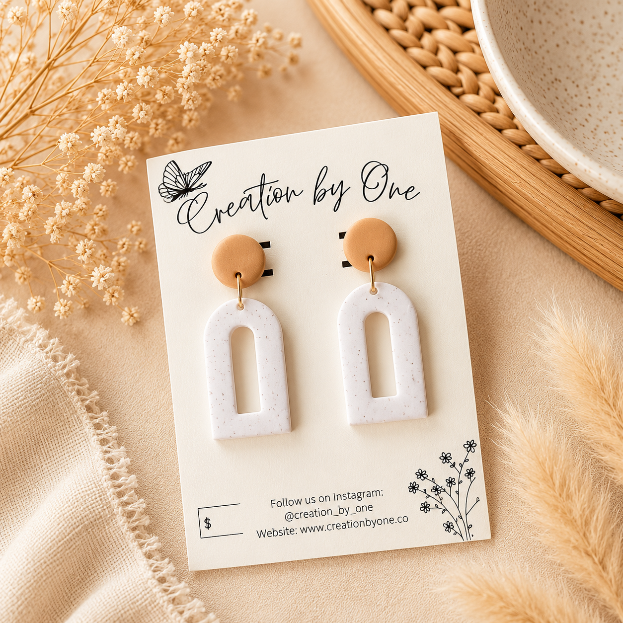 A pair of white speckled arch-shaped drop earrings with tan circular studs, displayed on a cream card labeled 'Creation by One' with dried flowers and a woven tray in the background.