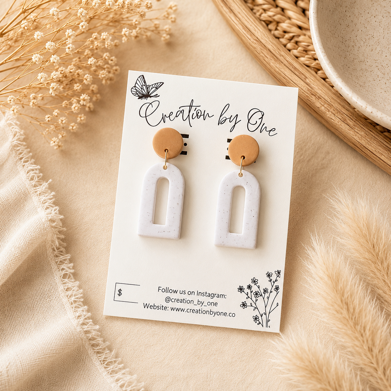 A pair of white arch-shaped drop earrings with round tan wooden tops, displayed on a white card labeled 'Creation by One' with neutral beige props around them.