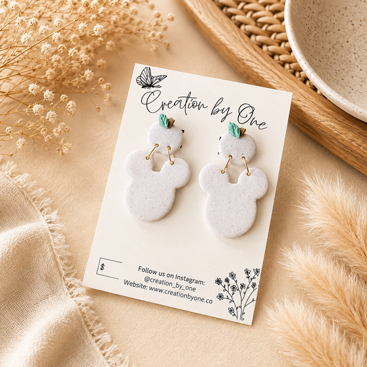 A pair of white ceramic snowflake-shaped dangle earrings with small gold hoops and mint green leaf accents, displayed on a white card labeled 'Creation by One' with dried floral props and a woven tray in the background.