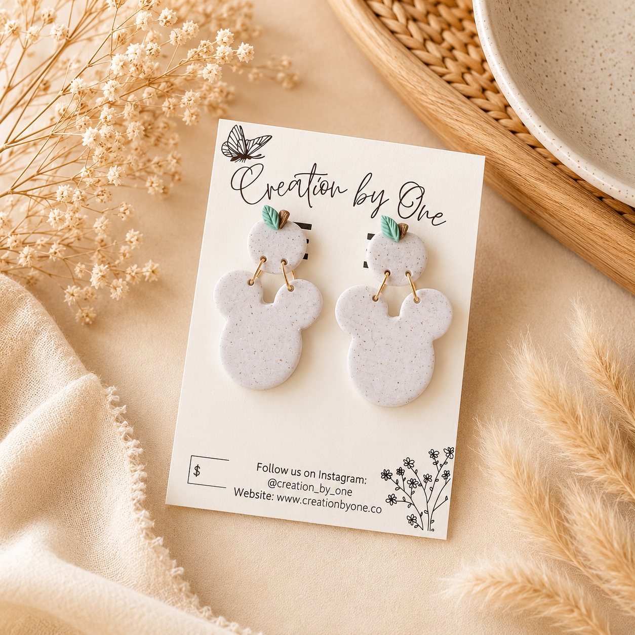 A pair of white speckled mouse-shaped earrings with small green leaf accents and gold rings, displayed on a branded card labeled 'Creation by One' against a soft beige fabric backdrop with dried flowers and a woven tray.
