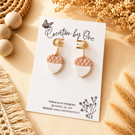 Pair of earrings on a 'Creation by One' branded card with decorative elements on a beige surface.