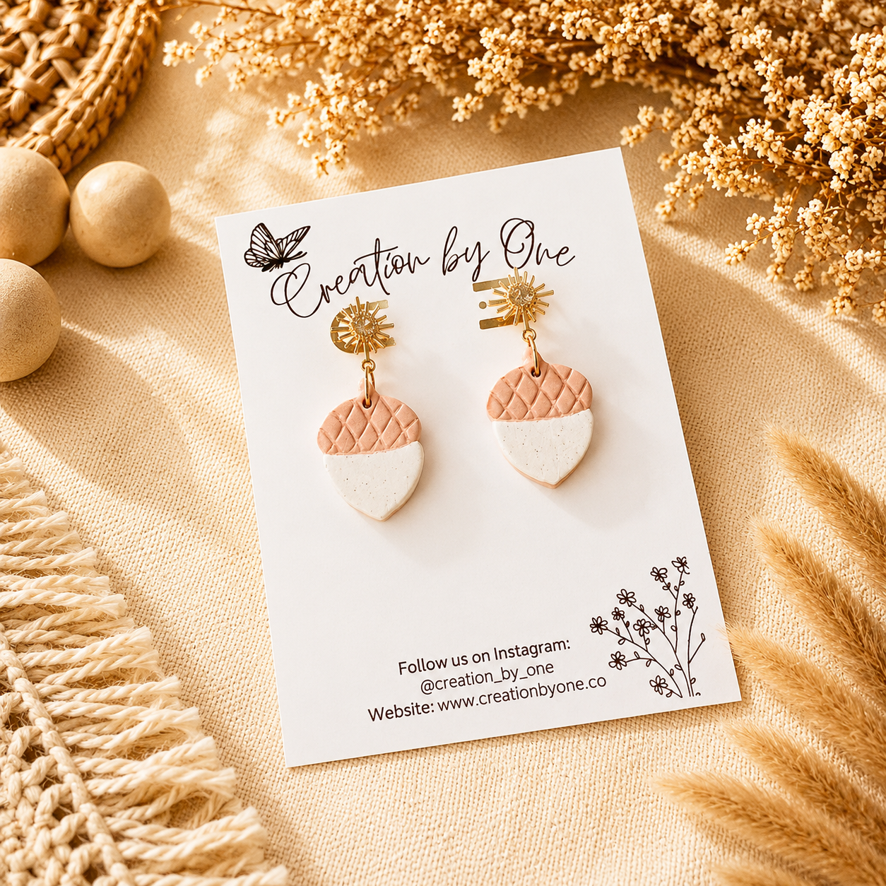 Pair of earrings on a 'Creation by One' branded card with decorative elements on a beige background.