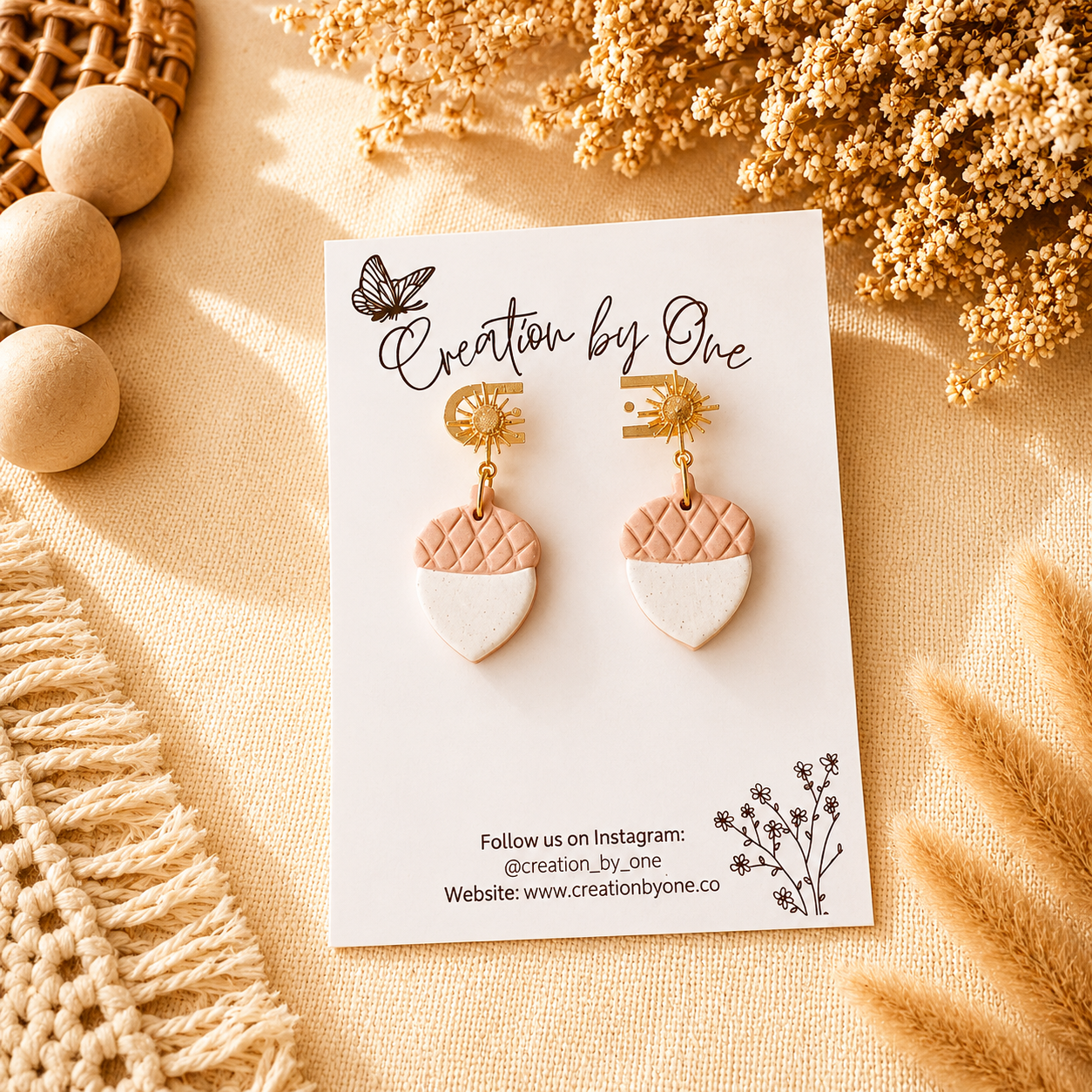 Pair of earrings on a 'Creation by One' branded card with decorative elements on a beige background.