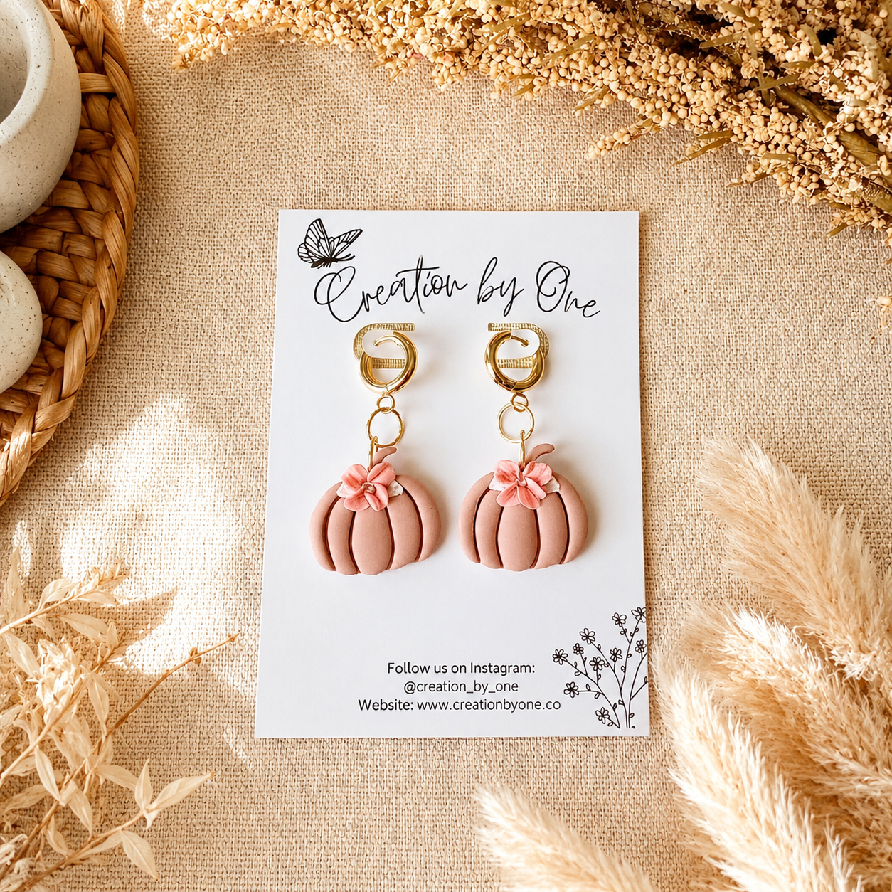 Pair of pink pumpkin-shaped earrings on a 'Creation by One' branded card with decorative elements.