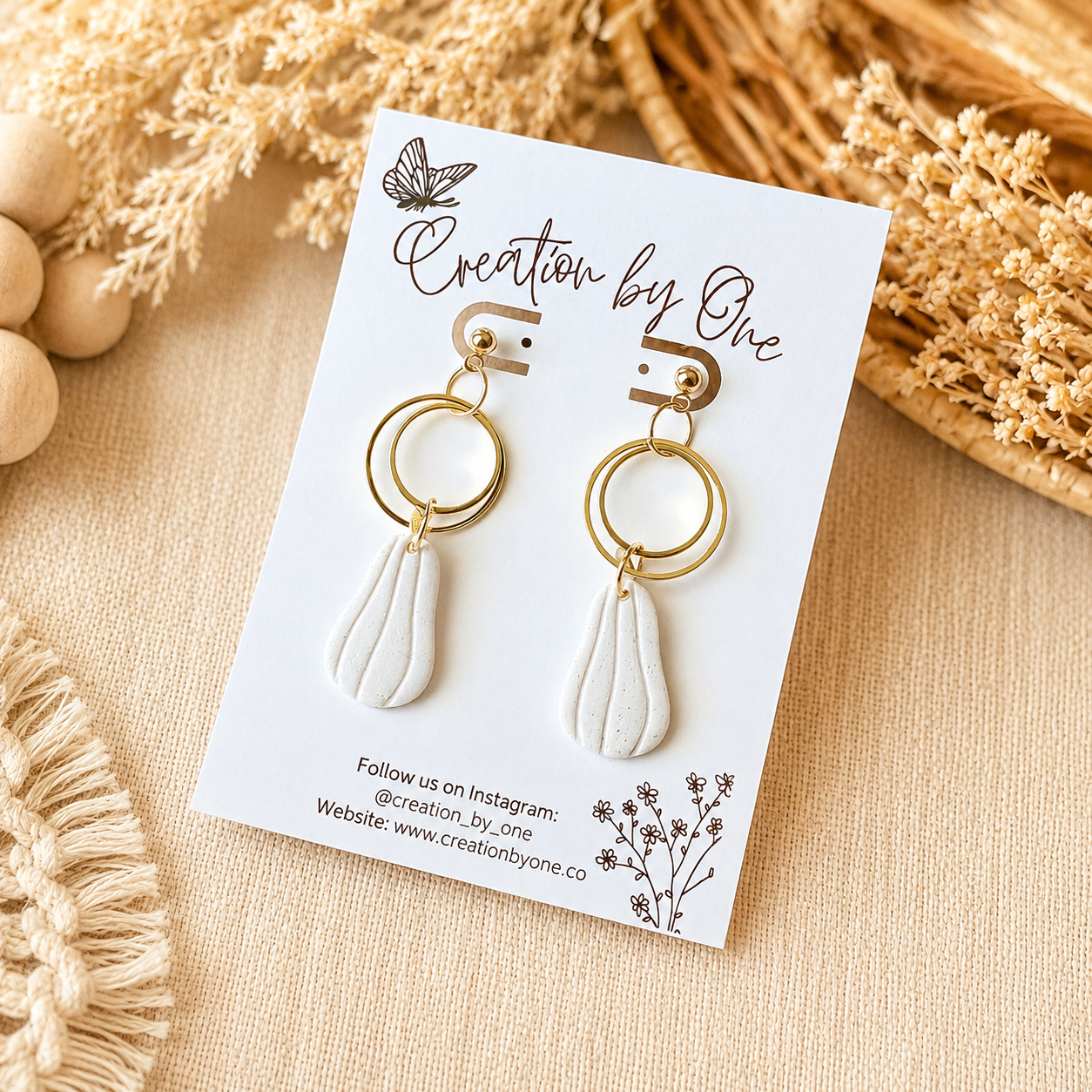 Gold and white earrings on a 'Creation by One' branded card with dried flowers in the background.