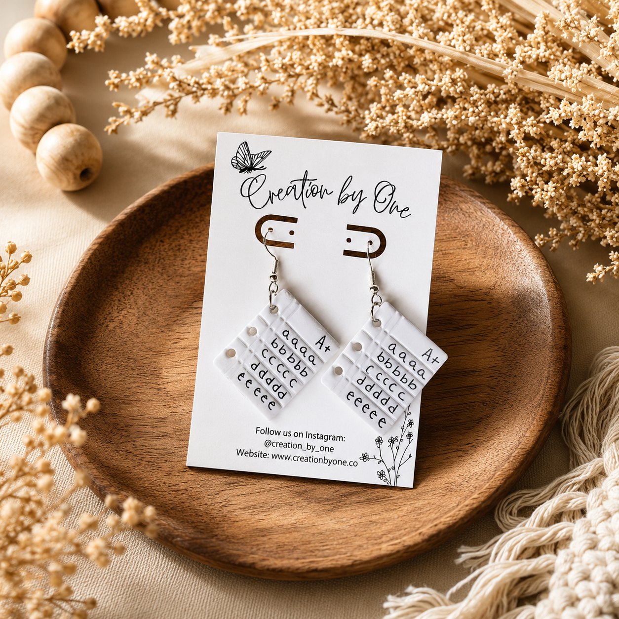 Pair of earrings on a 'Creation by One' card with decorative elements on a wooden plate.