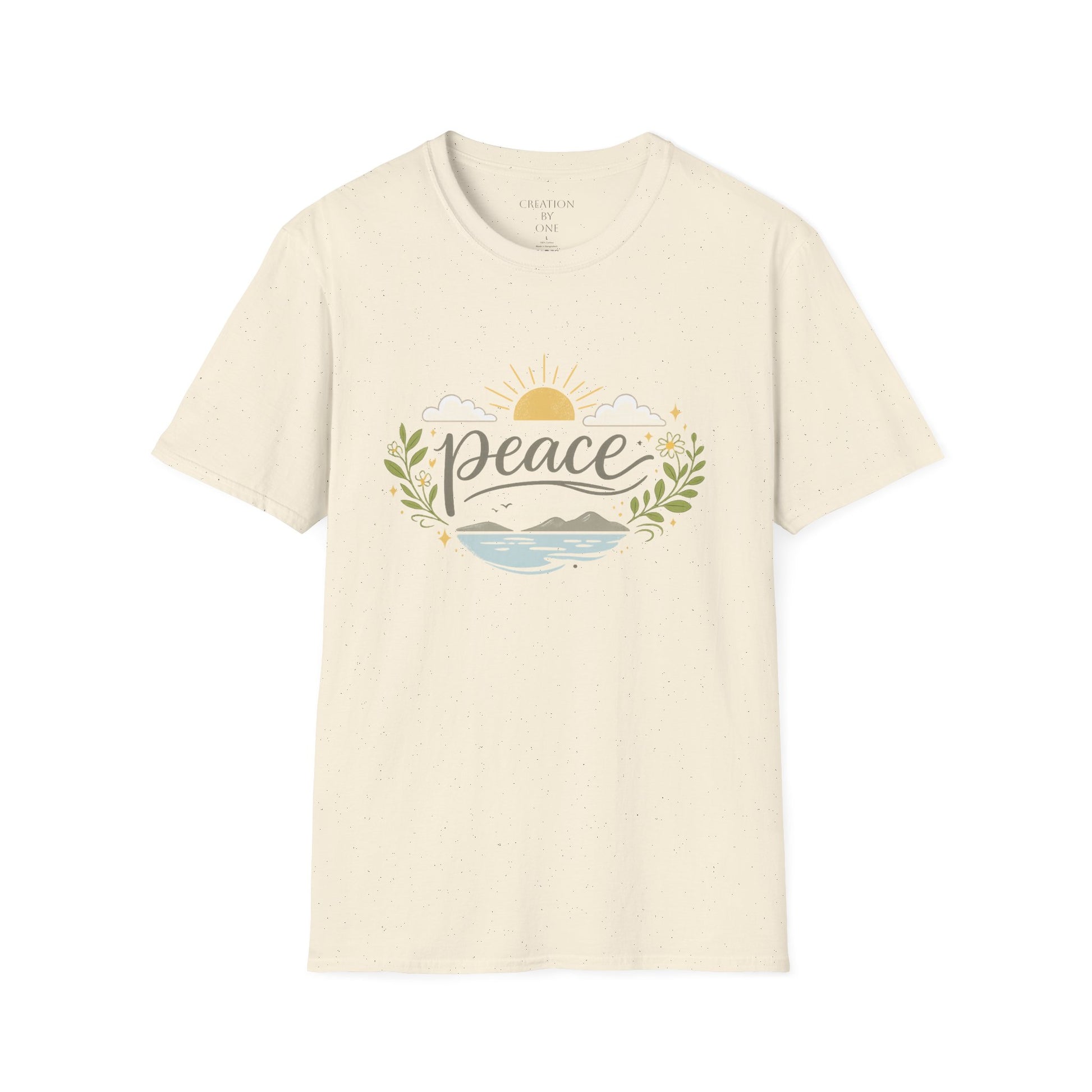 A woman with long dark wavy hair wears a neutral sand-colored graphic T-shirt and a beige baseball cap in a bright modern boho setting. The T-shirt features the word "Peace" in elegant script above a tranquil landscape with a golden sunrise, soft clouds, mountains, blue water, leafy branches, and delicate floral accents. She pairs the shirt with blue jeans and a simple gold cross necklace, standing in front of neutral décor with eucalyptus, pampas grass, and cozy textured accents for a calm, faith-inspired 