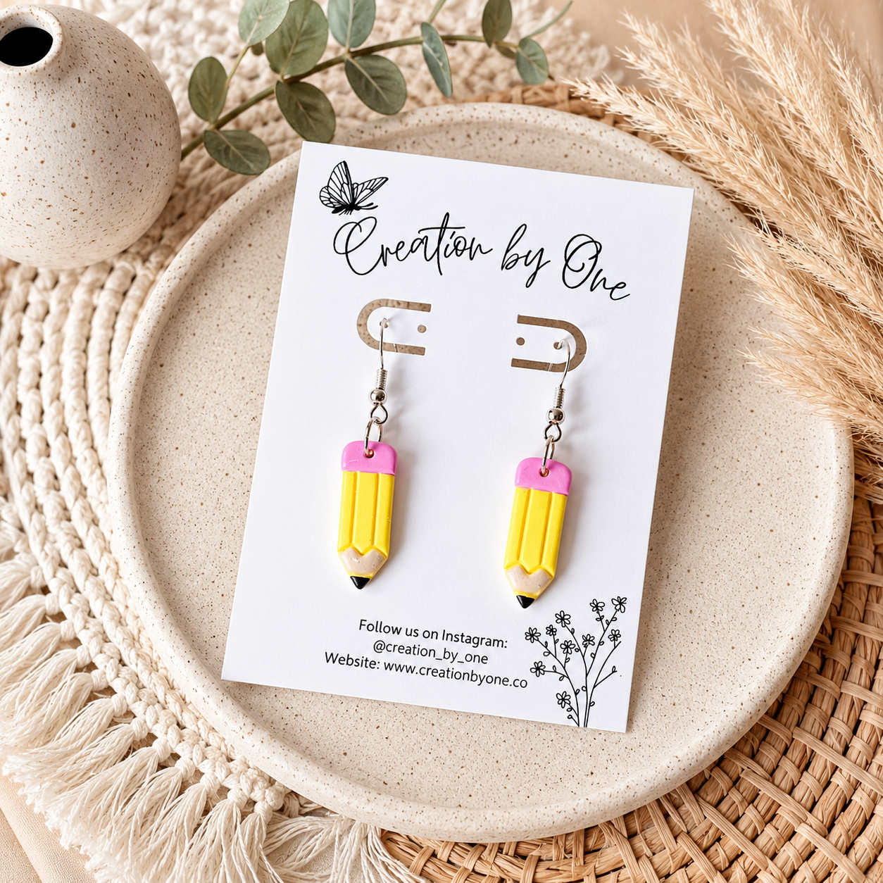 Yellow pencil-shaped earrings on a 'Creation by One' branded card, placed on a textured surface with decorative elements.