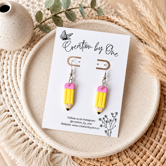 Yellow pencil-shaped earrings on a 'Creation by One' branded card, placed on a textured surface with decorative elements.