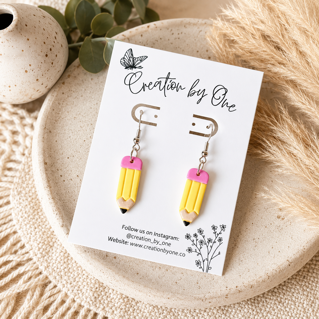 Yellow pencil-shaped earrings on a 'Creation by One' branded card, placed on a textured surface with a plant in the background.