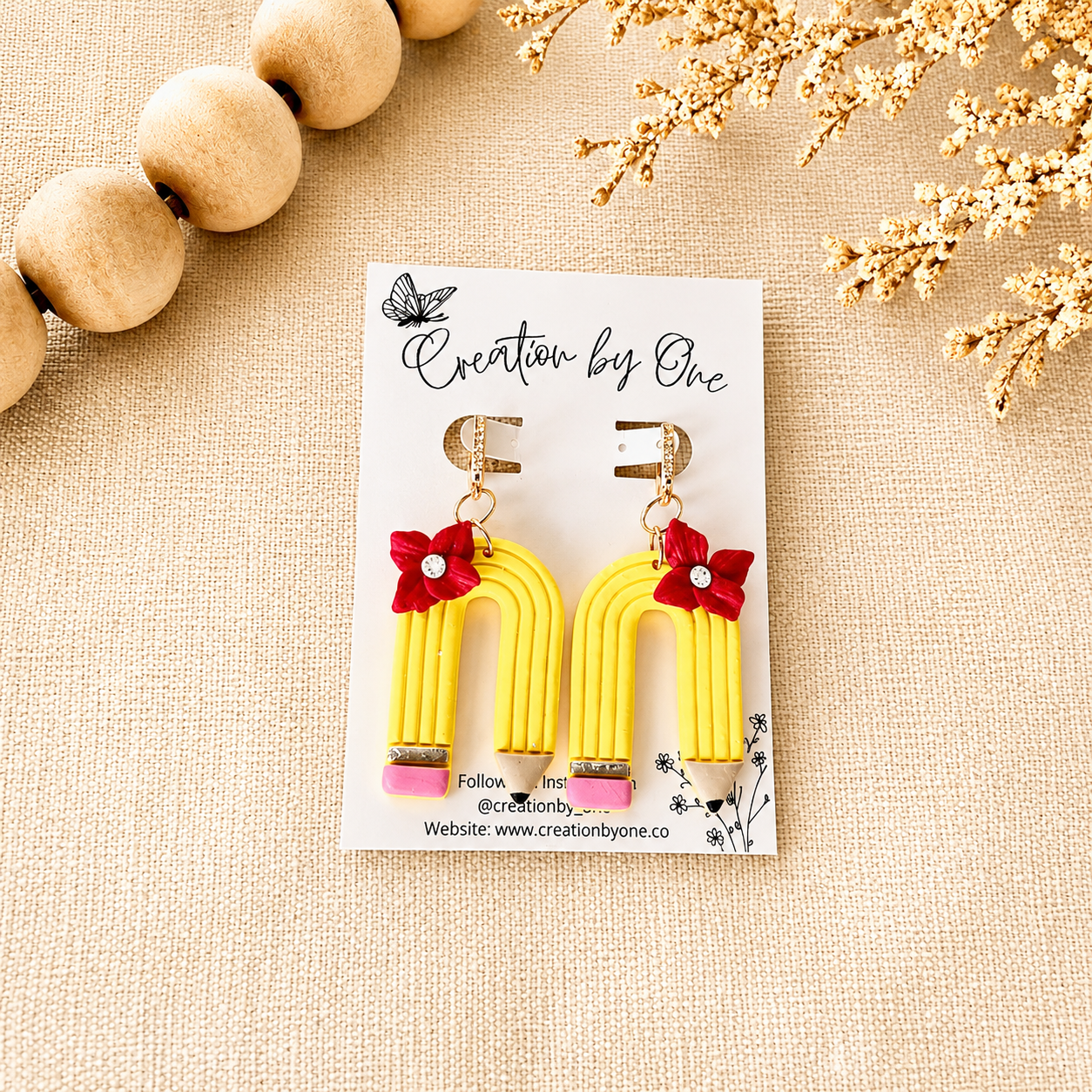 Yellow earrings with red flowers on a white card labeled 'Creation by One' on a beige background.