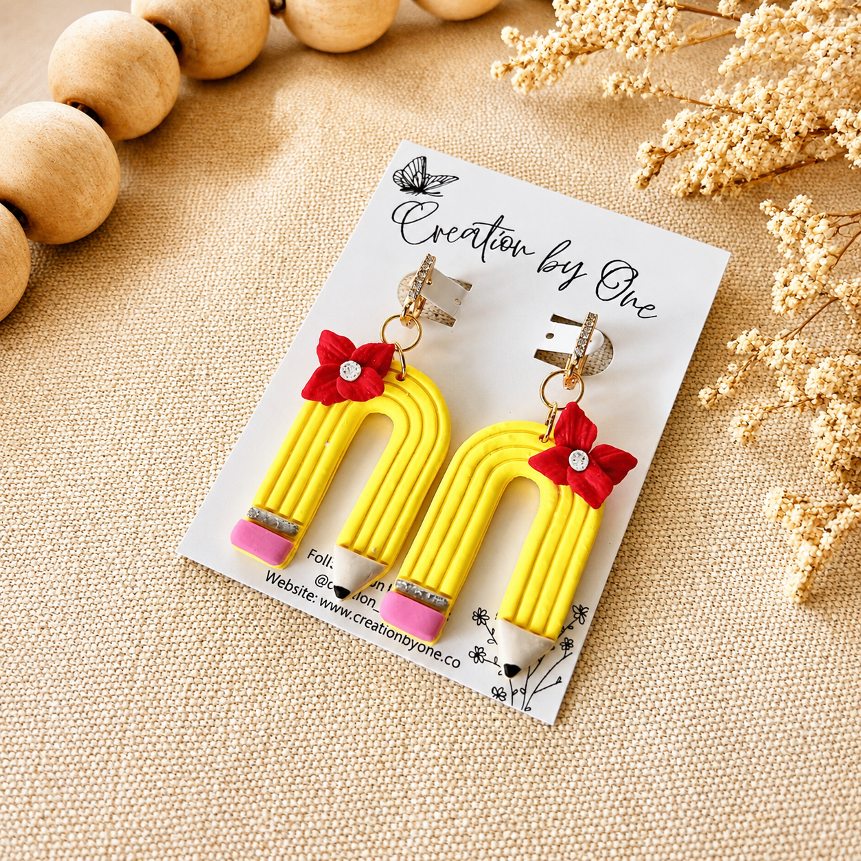 Yellow earrings with red bows on a 'Creation by One' card, placed on a textured surface.