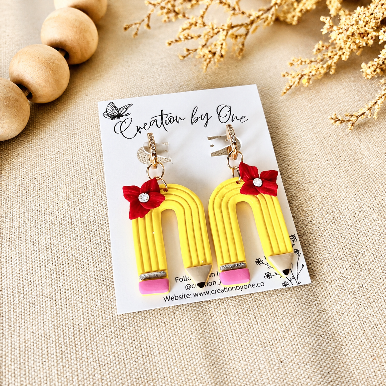Yellow pencil-shaped earrings with red bows on a 'Creation by One' branded card, placed on a textured surface.