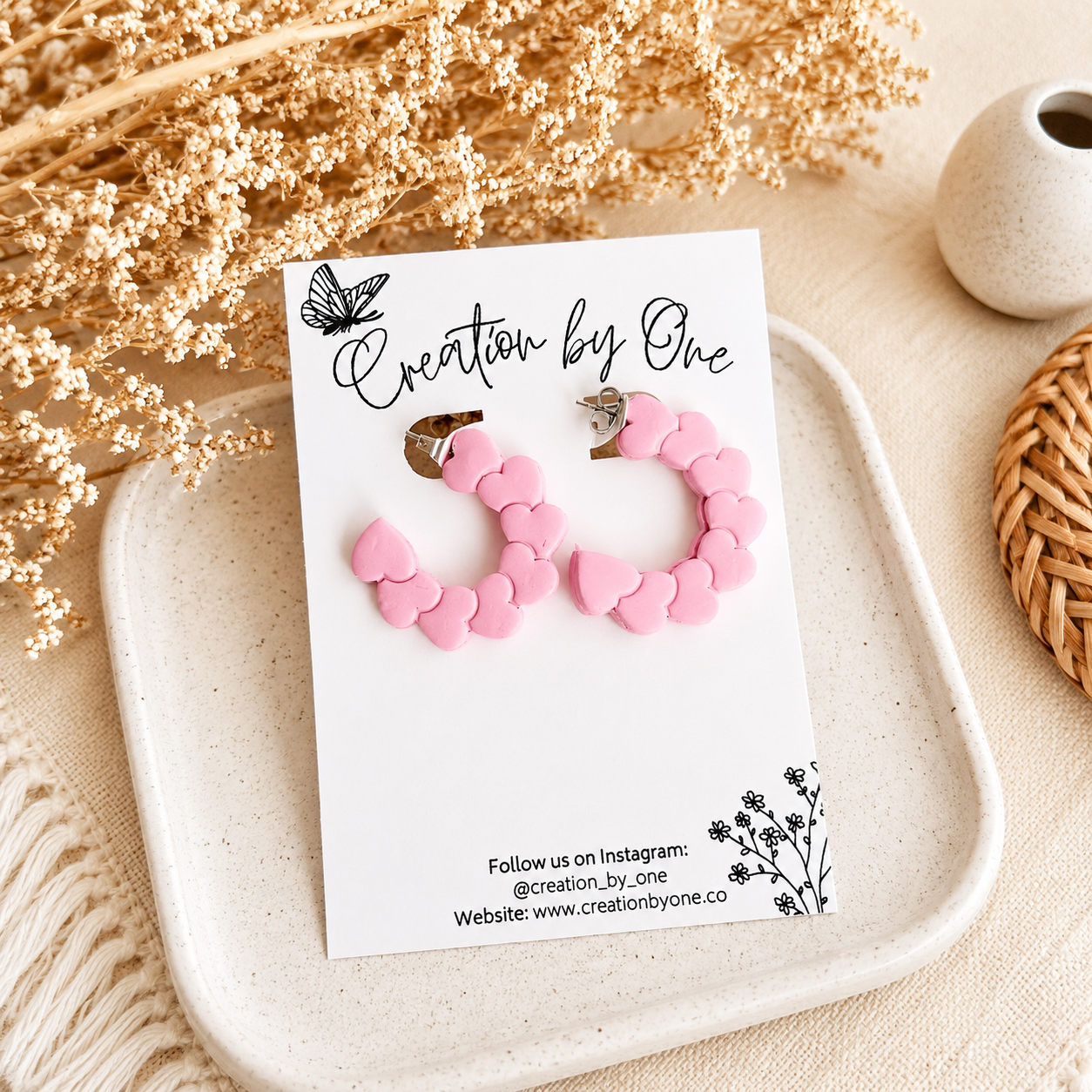 Pink heart hoop earrings on a Creation by One display card with a neutral boho background and dried floral accents.