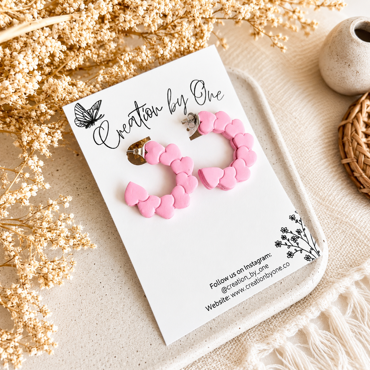 Pink heart hoop earrings on a Creation by One display card with a neutral boho background and dried floral accents.