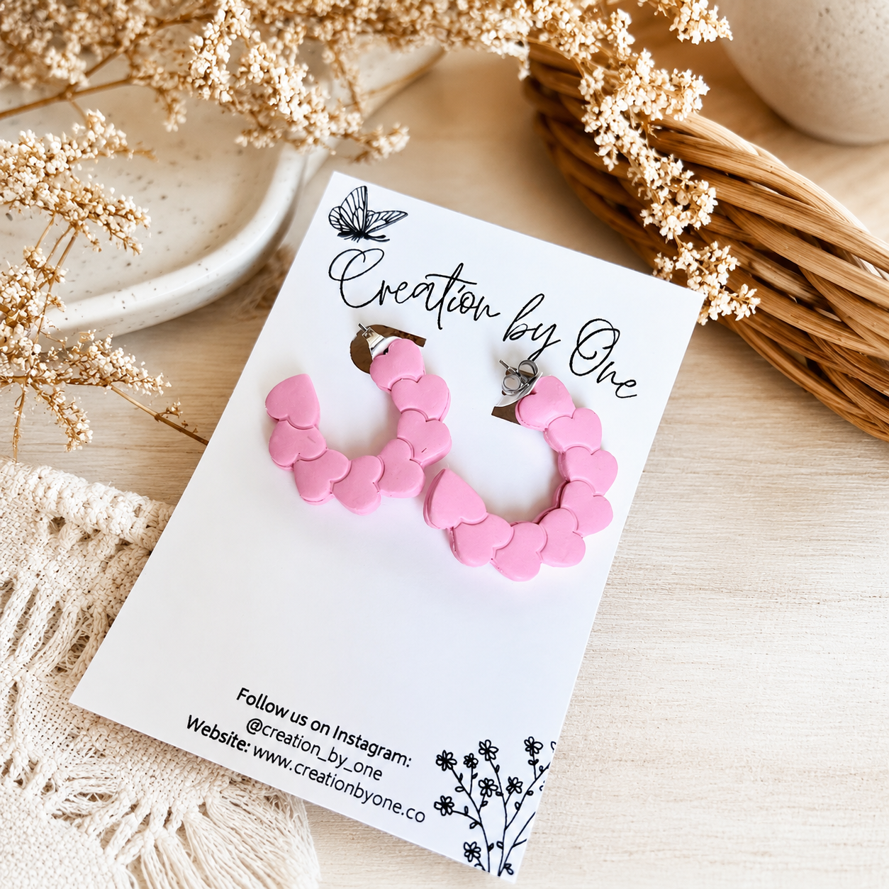 Pink heart hoop earrings on a Creation by One display card with a neutral boho background and dried floral accents.