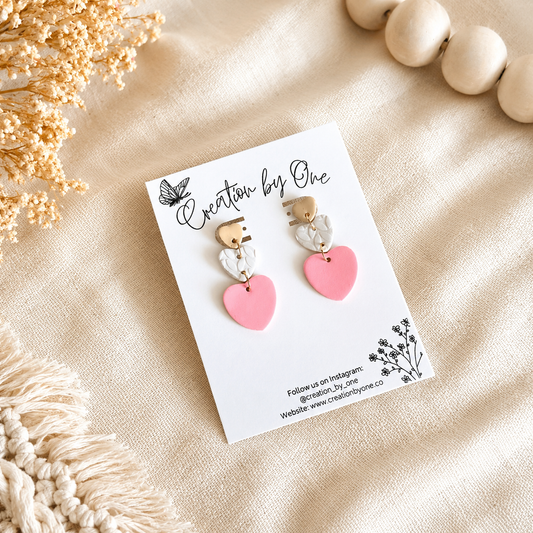 Handmade pink heart polymer clay dangle earrings displayed on a Creation by One jewelry card against a soft beige linen background with dried flowers, wooden beads, and boho décor in natural lighting.
