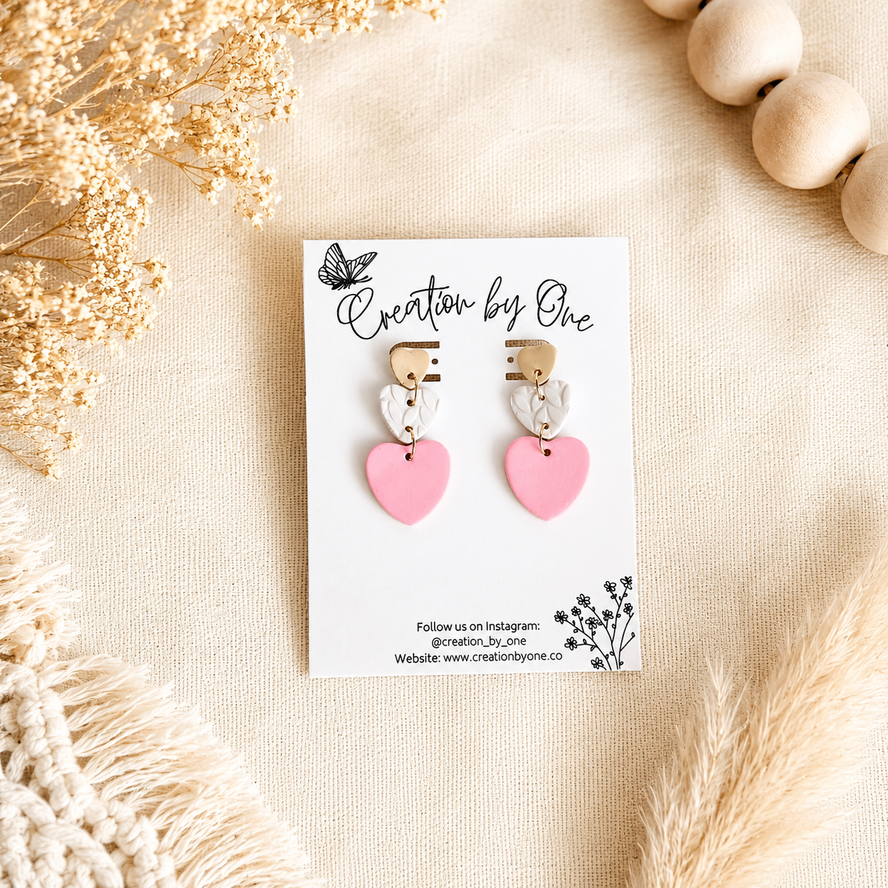 Handmade pink heart polymer clay dangle earrings displayed on a Creation by One jewelry card against a soft beige linen background with dried flowers, wooden beads, and boho décor in natural lighting.