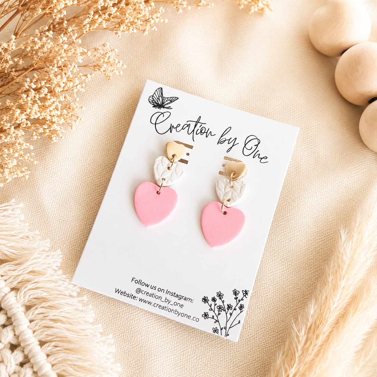 Handmade pink heart polymer clay dangle earrings displayed on a Creation by One jewelry card against a soft beige linen background with dried flowers, wooden beads, and boho décor in natural lighting.