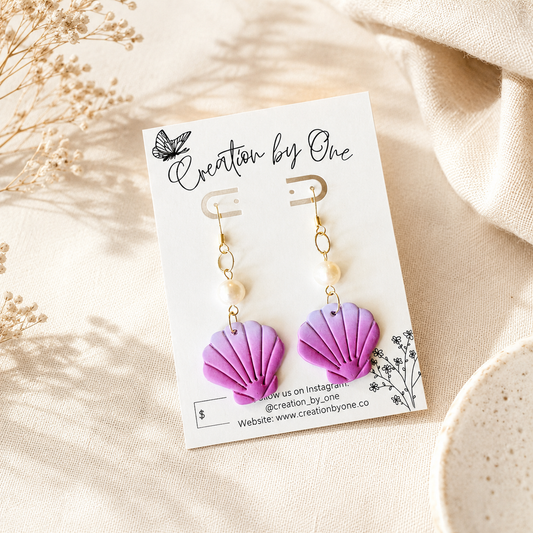 A pair of handmade shell-shaped drop earrings in purple and pink, hanging from gold hooks with white beads, displayed on a white card labeled 'Creation by One' on a beige fabric background.