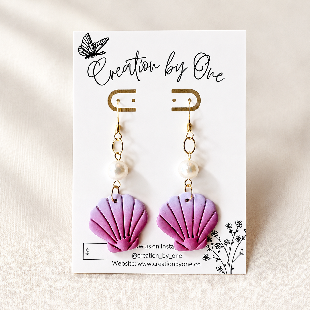 A pair of dangling shell-shaped earrings in pastel purple and pink, each with a small white pearl bead and gold hook, displayed on a white card labeled 'Creation by One' against a light fabric background.