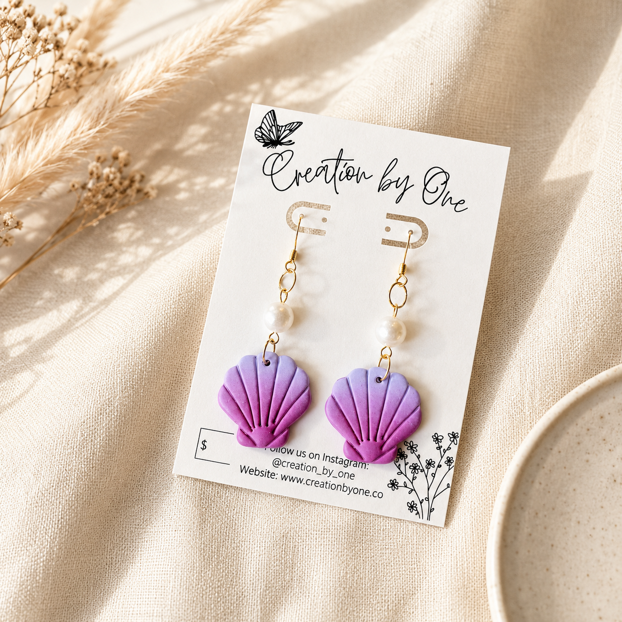 A pair of handmade drop earrings with purple and lavender shell-shaped charms, white pearl beads, and gold hooks, displayed on a white card labeled 'Creation by One' on a beige fabric background with dried grass and a ceramic plate.
