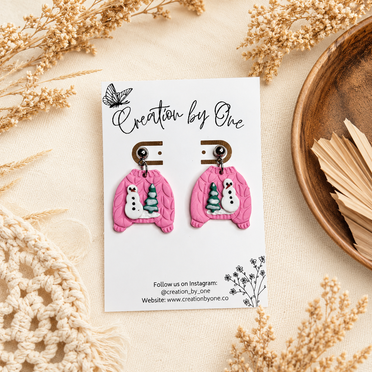 Handmade pink polymer clay dangle earrings shaped like cozy knit sweaters with snowman and Christmas tree details, displayed on a Creation by One jewelry card against a neutral boho linen background with dried florals.