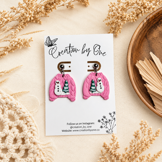 Handmade pink polymer clay dangle earrings shaped like cozy knit sweaters with snowman and Christmas tree details, displayed on a Creation by One jewelry card against a neutral boho linen background with dried florals.