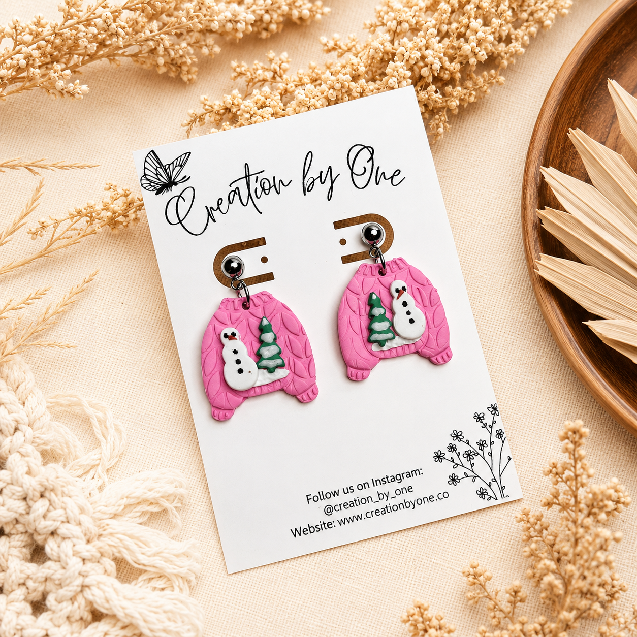 Handmade pink polymer clay dangle earrings shaped like cozy knit sweaters with snowman and Christmas tree details, displayed on a Creation by One jewelry card against a neutral boho linen background with dried florals.