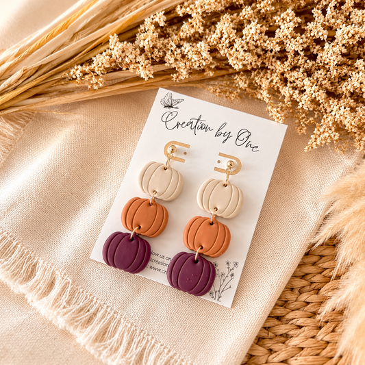 Pair of earrings on a card with 'Creation by One' branding, displayed on a textured surface.