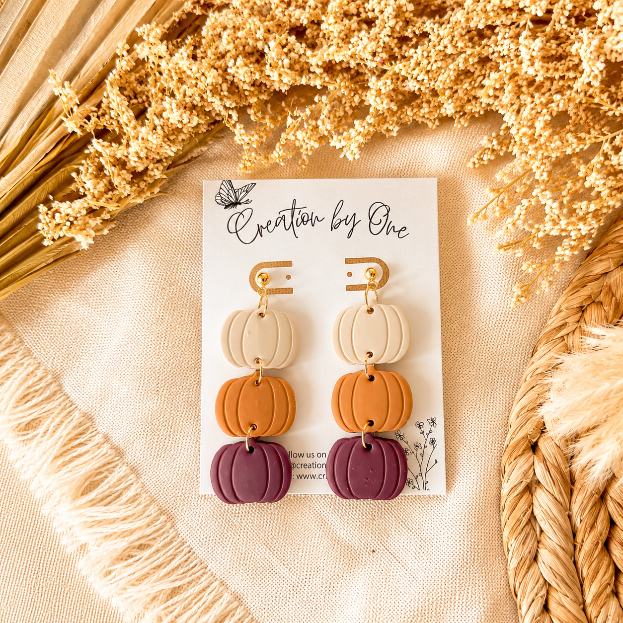 Pair of pumpkin-shaped earrings on a 'Creation by One' card with dried flowers and woven elements in the background.