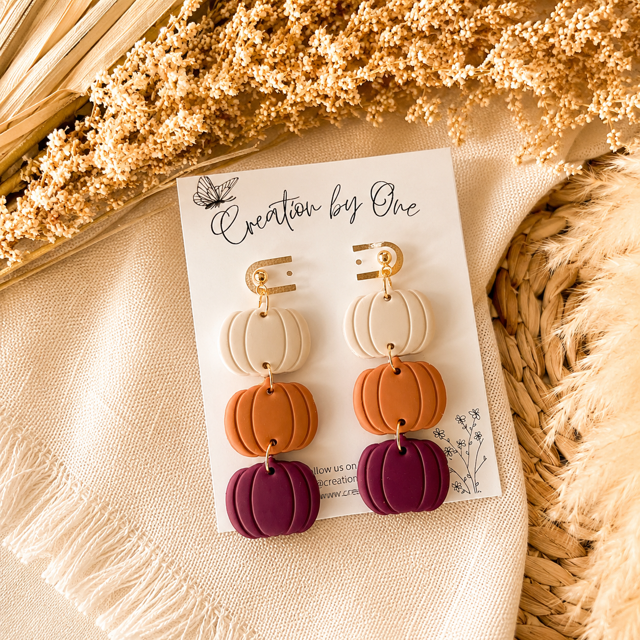 Colorful pumpkin-shaped earrings on a 'Creation by One' branded card with dried flowers and woven material in the background.
